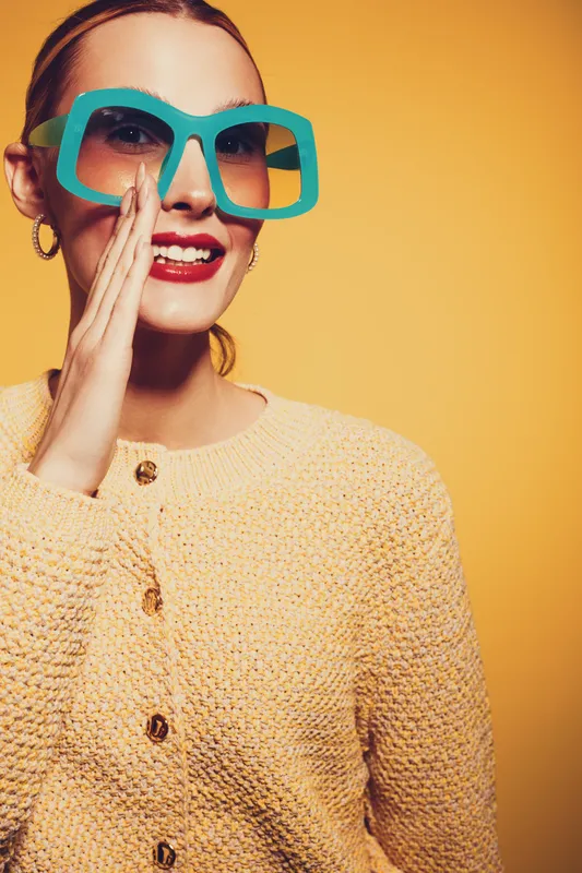 Smiling woman in oversized turquoise sunglasses and yellow knitted sweater poses against yellow background. Fashion portrait with bright colors and trendy accessories concept.