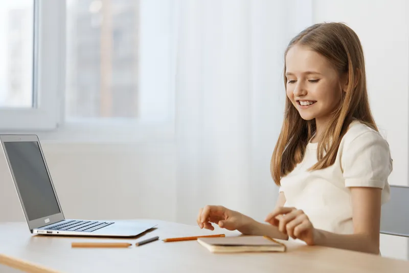 A cheerful young girl with long hair, sitting at a desk with a laptop, engaged in creative activities and smiling in a bright, minimalistic workspace