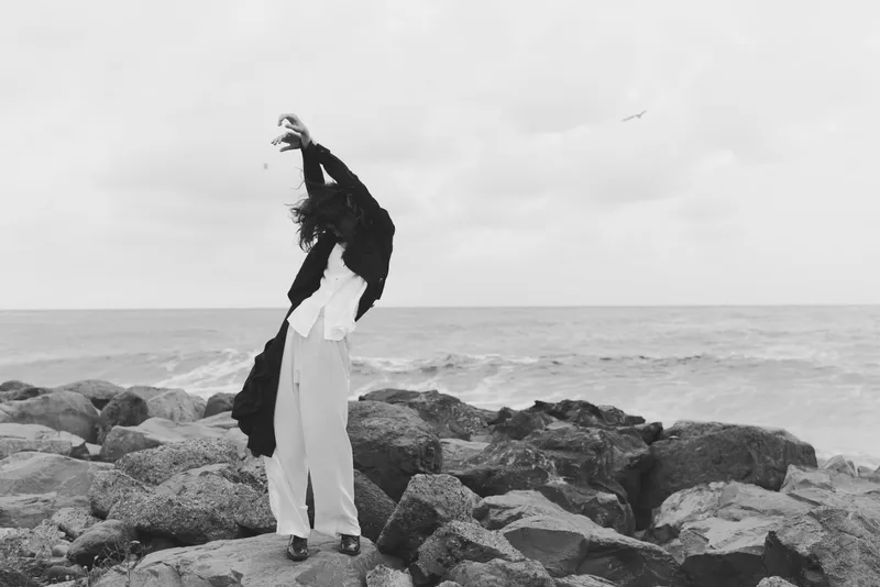 Monochrome shoreline scene features a figure on rugged rocks with arms raised, wind catching the coat, expansive ocean backdrop and a minimalist, dramatic coastal mood, evoking freedom and release.