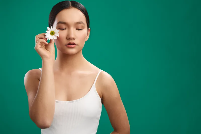 Asian woman stands against a vibrant green backdrop, gently holding a white flower near her eye with a calm, thoughtful expression, radiating serenity, natural beauty, and mindful poise in a clean