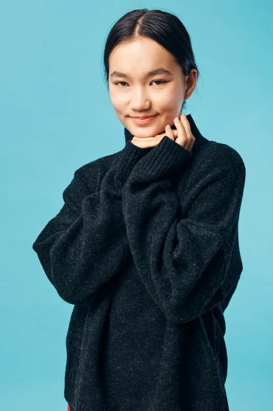 Young woman in black sweater smiling and posing with hands near face on blue background. Asian female with natural makeup and casual outfit in studio for portrait photography.
