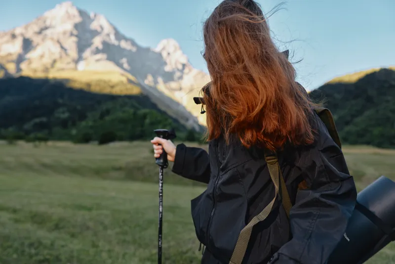 A female hiker with a backpack stands in a wide valley, gazing toward distant peaks while capturing the moment with a camera amid a rugged, sunlit landscape.