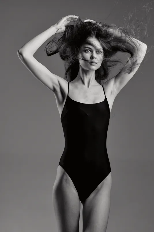 Black and white portrait of a slim woman in a black bodysuit with flowing hair and serious expression against gray background