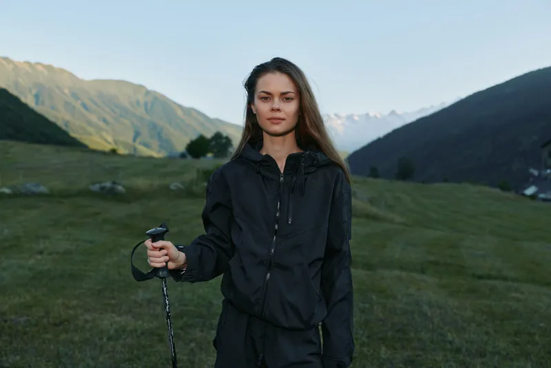 A young woman in a black outdoor jacket stands in a valley with expansive mountains behind. She grips a trekking pole, looks at the camera with calm confidence, evoking adventure and resilience.