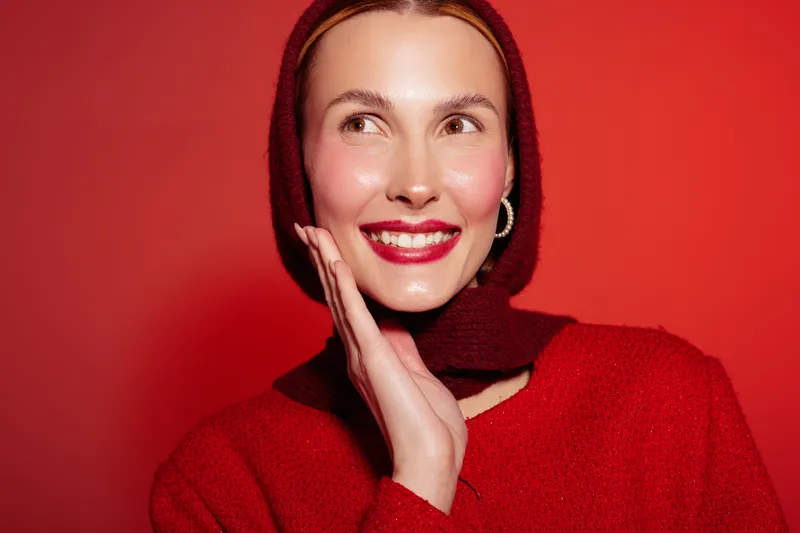 Smiling woman in red sweater and headscarf poses against red background, showing bright makeup and joyful expression. Concept of fashion, beauty, and vibrant style.