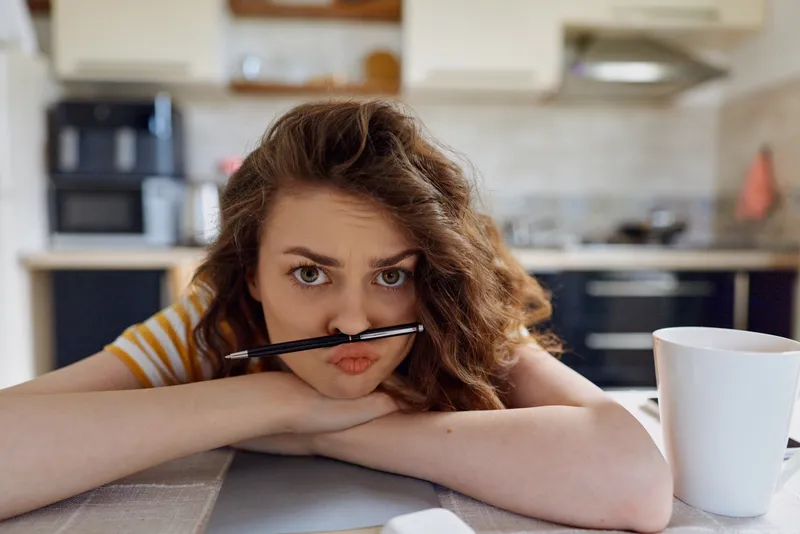 Young woman rests her chin on folded arms at a kitchen island, holding a pen to her lips in a bored contemplative moment during a quiet home morning.