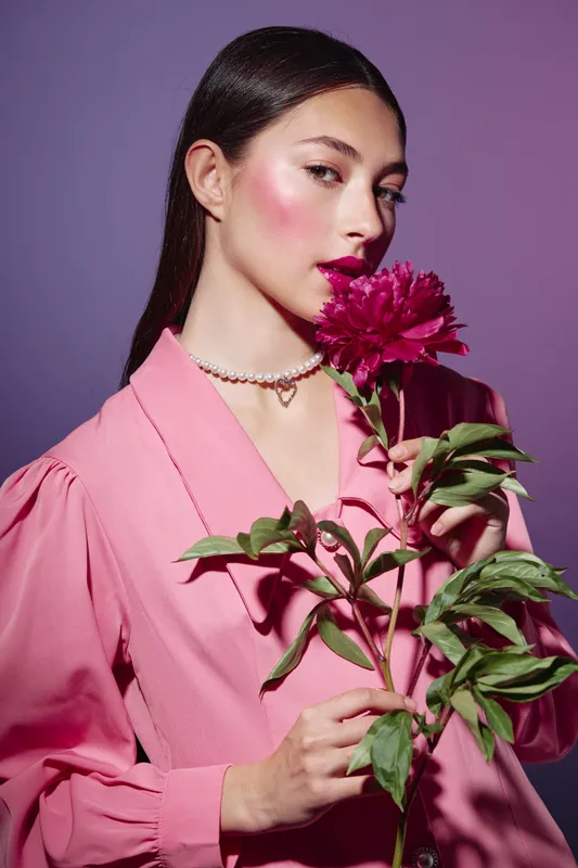 Young woman in a pink outfit holds a vibrant flower near her face, showcasing creative beauty and fashion with glossy makeup and elegant accessories on purple background.