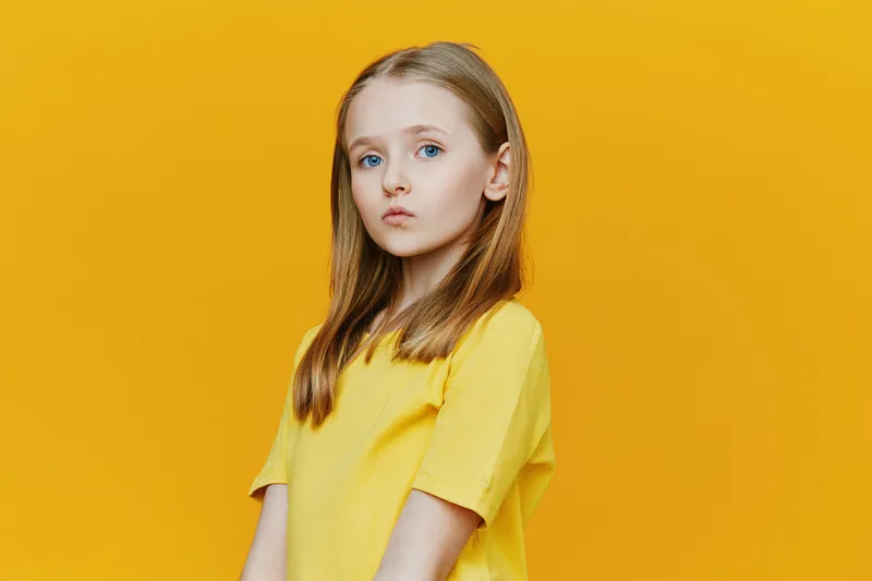 A young girl with long blonde hair stands against a bright yellow background, wearing a yellow shirt, embodying confidence and innocence with a serious expression