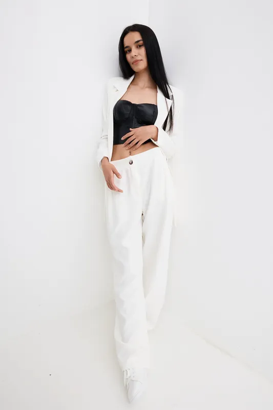 Stylish woman in white pants and black bustier poses confidently against a plain background, showcasing modern fashion and minimalistic elegance in a studio setting.