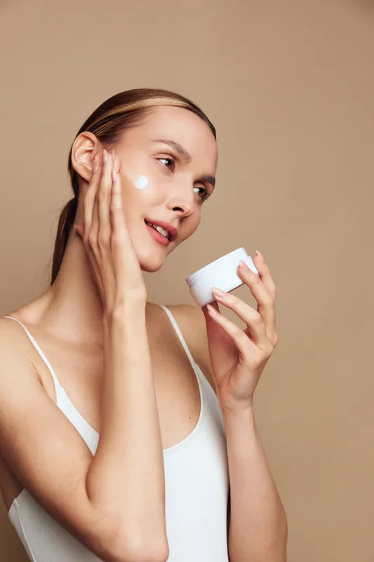 Woman skincare beauty portrait, applying moisturizer cream to face with fingers, holding small jar, calm expression, studio background, soft light, close up, beauty care routine, skin care
