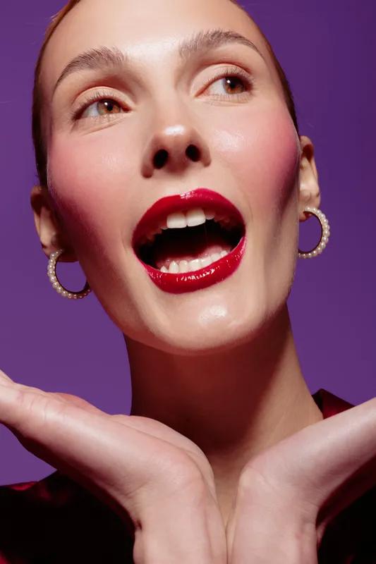 Close-up of a woman with glossy red lips and pearl earrings posing with hands under chin on purple background. Concept of beauty, makeup, fashion, and elegance.