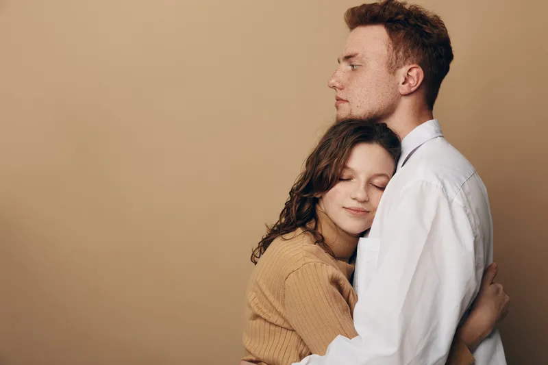 Young couple embraces tenderly with closed eyes, expressing love and comfort. The woman wears a brown sweater, the man a white shirt, against a plain beige background.