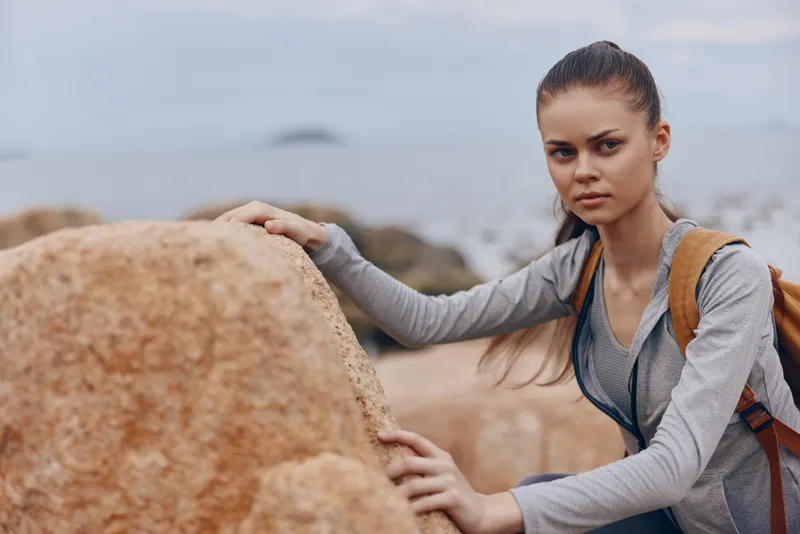 Young female hiker exploring rocky shoreline with serious expression, wearing sporty outfit and backpack against sea backdrop, perfect for adventure and outdoor themes