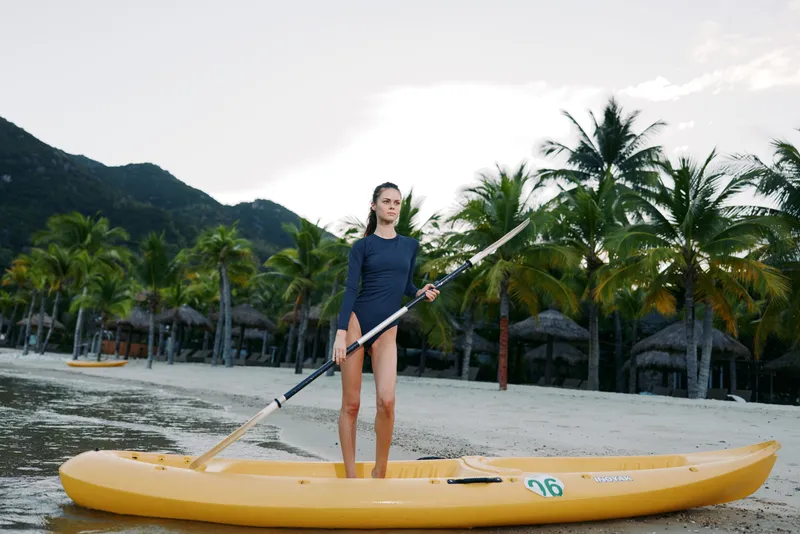 Young woman stands on a bright yellow paddleboard, gripping a paddle on calm water, framed by palm trees and a sandy shore with distant mountains under a soft sky.