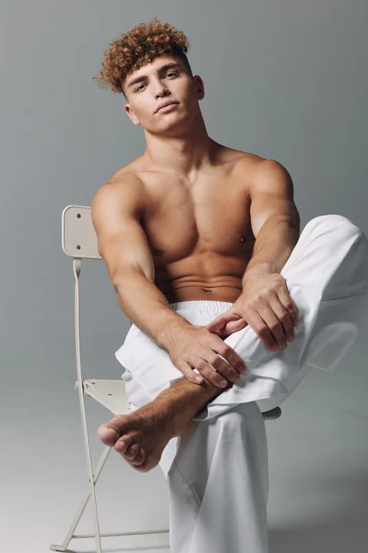 Young man with curly hair sitting shirtless on chair in studio, relaxed pose with white pants and neutral background for fashion or lifestyle concept.