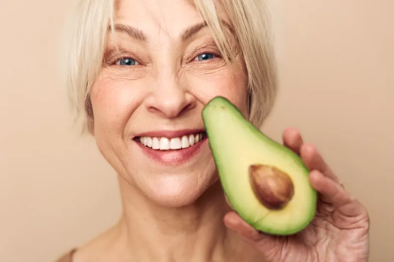 Smiling mature woman with short blonde hair holds fresh avocado half near face. Healthy lifestyle, nutrition concept with natural fruit, wellness, and positive mood on beige background.