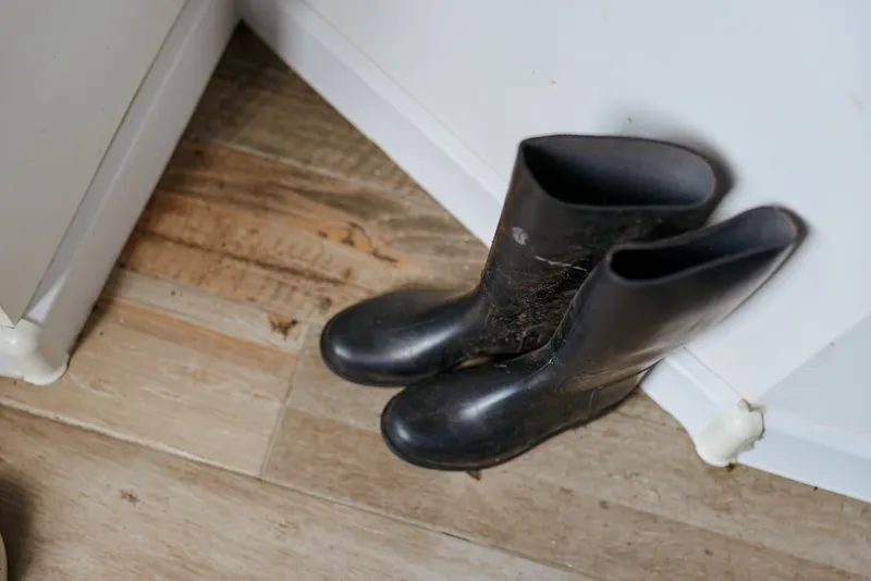 Casual indoor scene showing boots resting by a wall in a home, a simple lifestyle moment with rustic wood floor, natural lighting, calm atmosphere, minimal decor, everyday detail, intimate domestic
