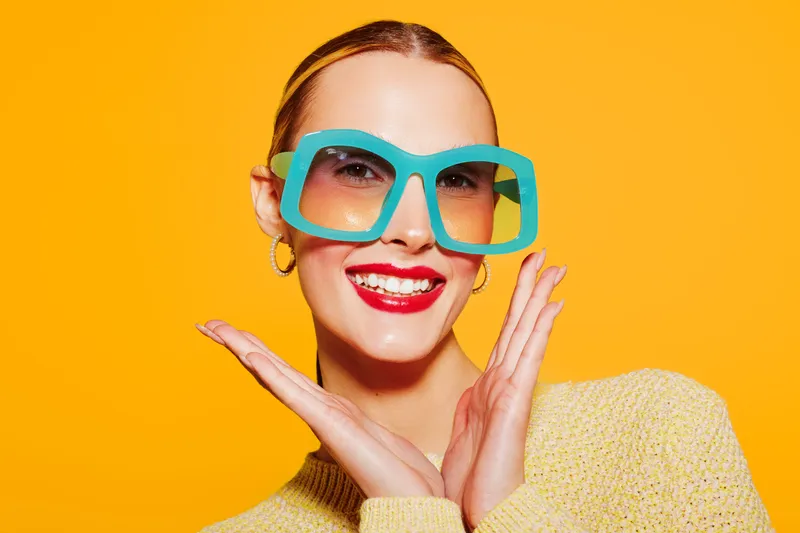 Smiling woman with large blue sunglasses and red lipstick poses against bright yellow background. She wears a yellow sweater and gold hoop earrings. Concept of fashion, style, and happiness.