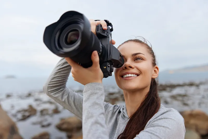 Young woman capturing moments with a camera on the beach, showcasing a joyful emotion while dressed in casual grey clothing against a serene coastal background