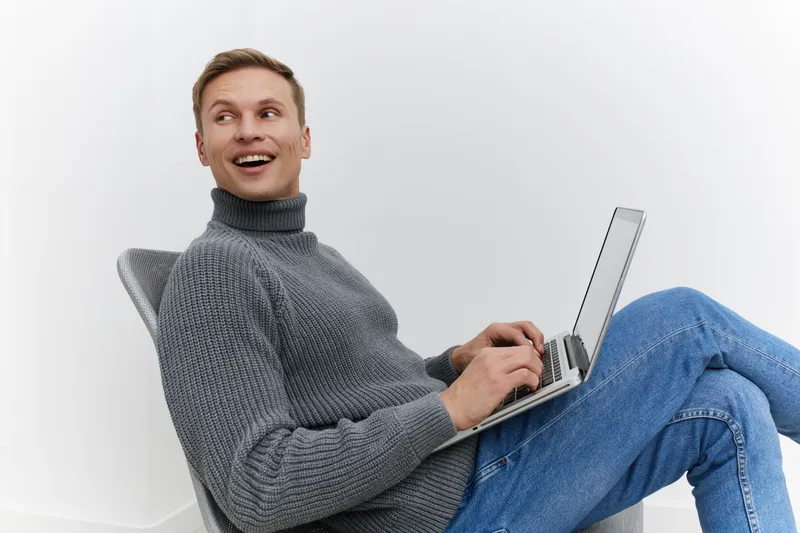 Happy smiling man with short brown hair, casual gray turtleneck and jeans sitting on modern chair, working on laptop, isolated on plain white background, studio portrait, people lifestyle concept, mod