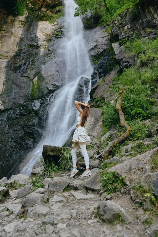 woman hiking near waterfall in forest, wearing casual clothes and sneakers, surrounded by rocks and greenery, outdoor adventure and nature exploring