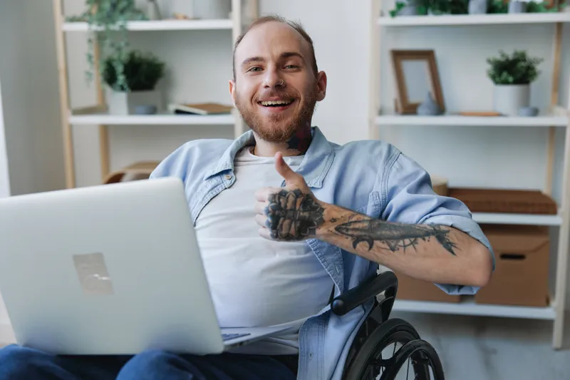 Мужчина в инвалидной коляске, фрилансер, улыбаясь работает за ноутбуком дома