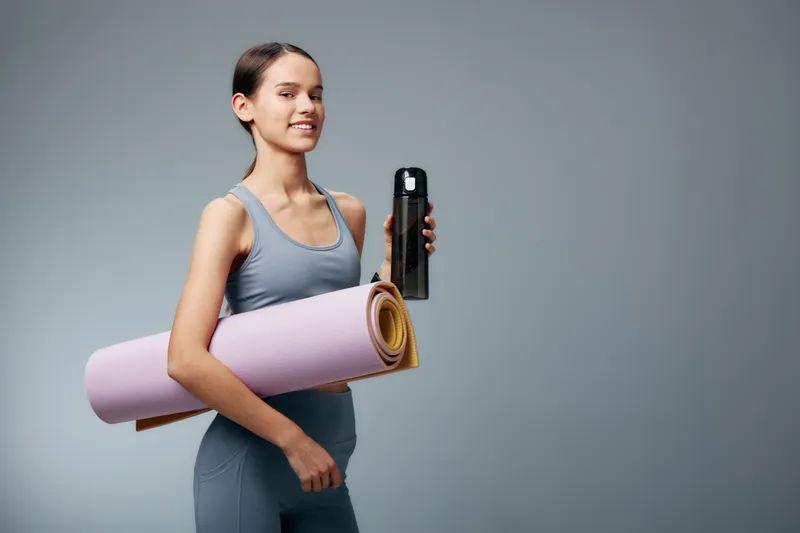 Fitness woman in athletic wear carries a yoga mat and water bottle, ready for a focused workout session in a clean minimalist studio, conveying health and discipline.