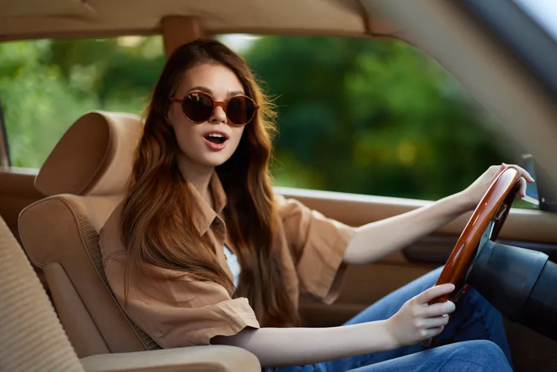Young woman with long hair driving a vintage car, wearing stylish sunglasses and a casual shirt, expressing excitement in a lush green background.