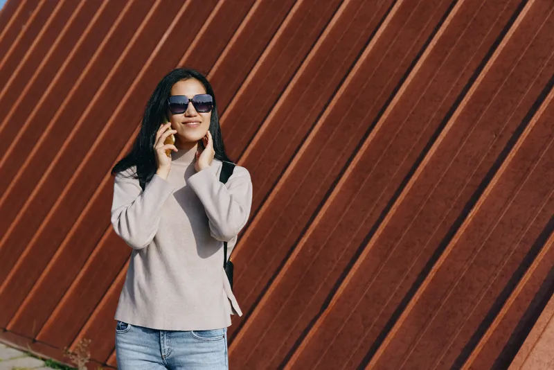 Woman wearing sunglasses talks on mobile outdoors against a diagonal wooden wall, casual sweater and denim, sunlit street scene, stylish, relaxed lifestyle shot with modern urban vibe.