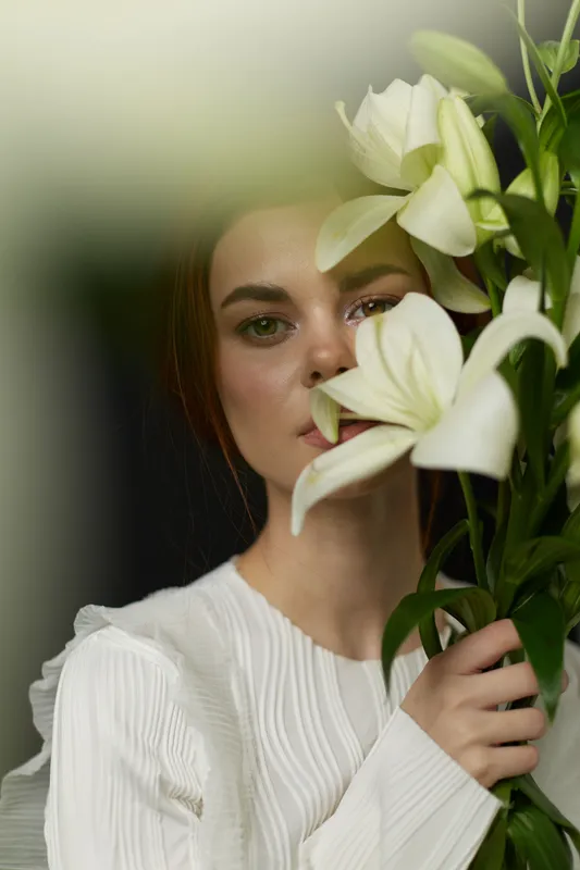Young woman with long dark hair holding white lilies, showcasing serene emotion, dressed in a textured white blouse, against a soft focus background