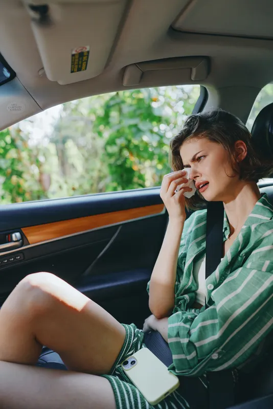 A woman sits in the front passenger seat of a car, looking distressed and wiping tears while resting her head.
