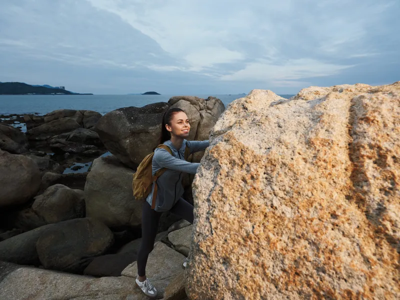 A young woman of Asian descent climbs over a large rock on a rugged beach, demonstrating determination and adventure in a stunning coastal setting