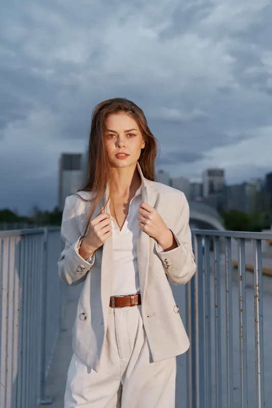Confident woman in business attire standing outdoors against a dramatic skyline, showcasing modern elegance and professionalism, with a neutral color palette.