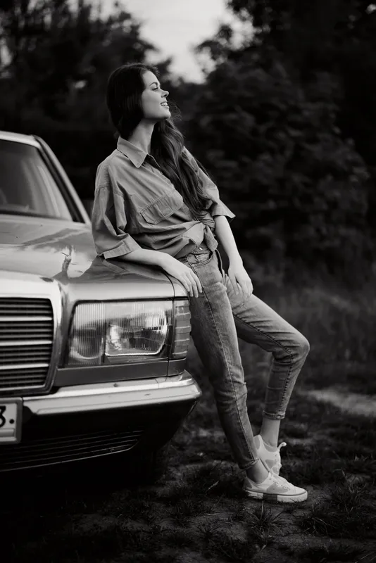 Stylish woman in casual attire leaning on vintage car, evoking freedom and nostalgia, captured in black and white photography.