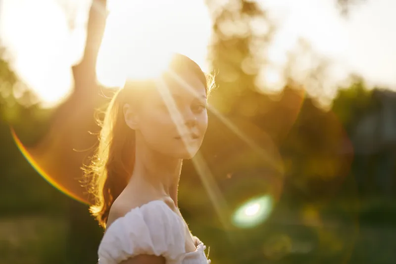 Young woman in a white dress smiling at sunset, embodying joy and serenity in nature with warm light and soft bokeh effect