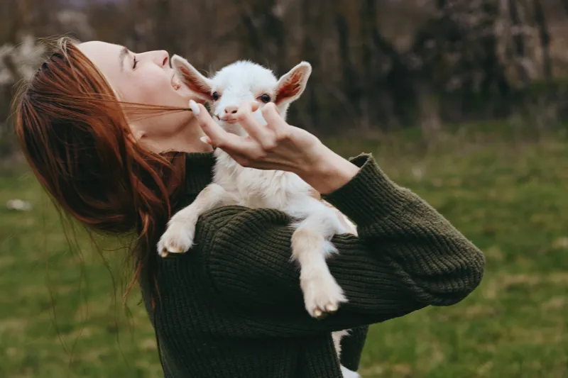 Cheerful woman outdoors, holding a goat kid close, sunlit moment, relaxed posture, soft breeze, natural colors, rustic setting, gentle laughter, intimate bond, countryside mood