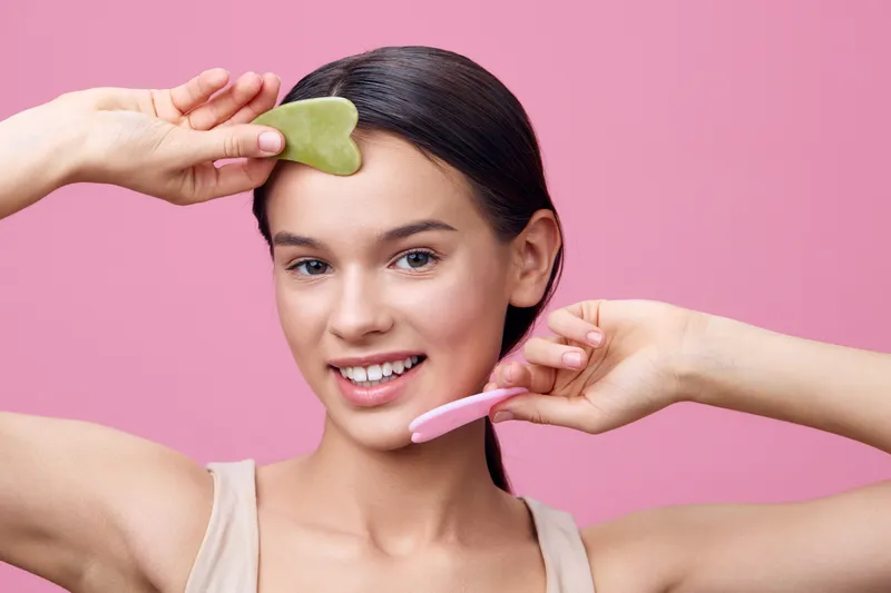 Young woman using a gua sha stone on her face, smiling against a soft pink background, embodying self care and beauty concepts with a joyful expression