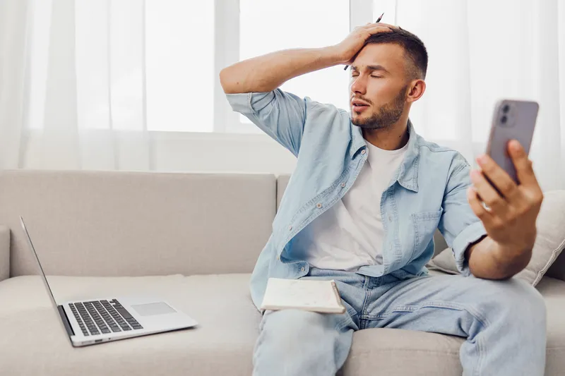 Stressed man sitting on a couch, holding a phone and a notebook, surrounded by a cozy, well-lit living space. He expresses deep concentration and frustration.
