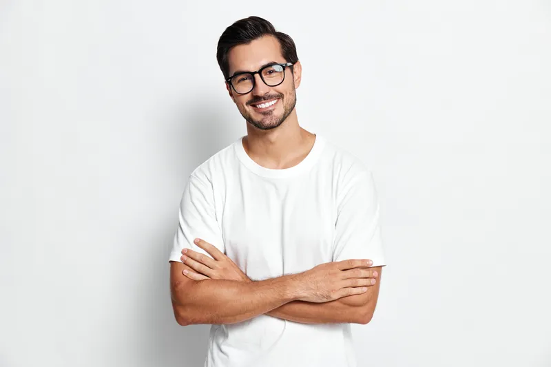 Smiling man with glasses in a white t-shirt stands confidently with crossed arms against a plain light background. Professional portrait in casual style.