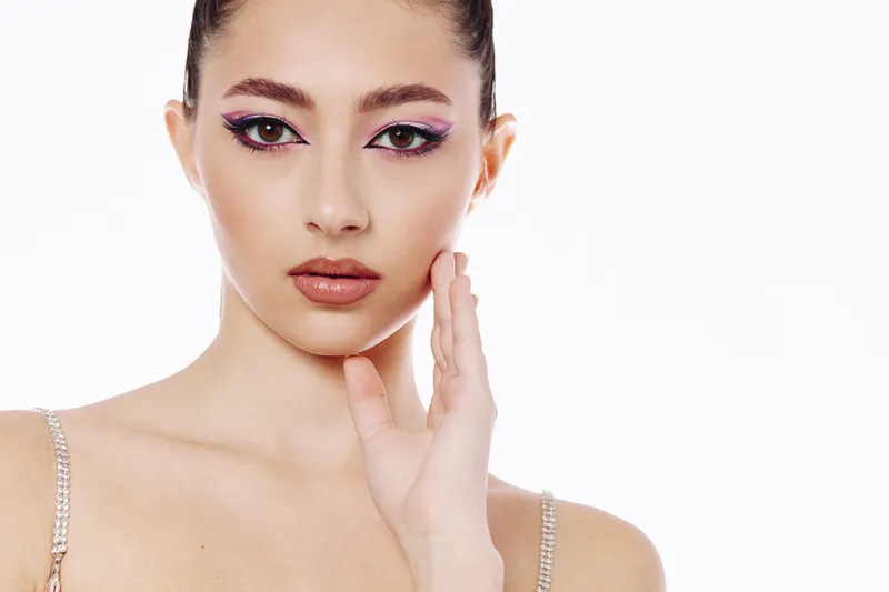 Young woman with vibrant creative makeup and glossy lips poses against a white background. Detailed facial features highlight modern beauty and fashion concepts, new trends.