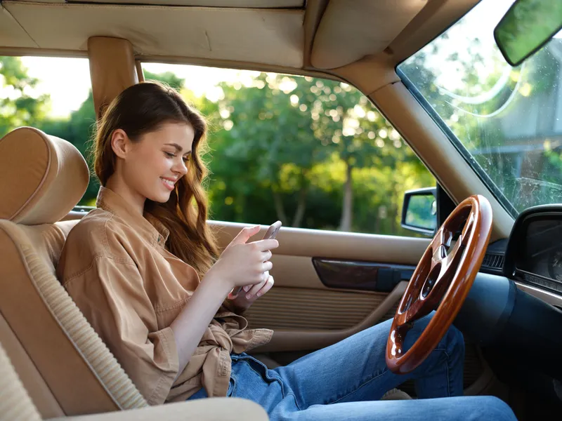 Young woman smiling while using smartphone in a vintage car interior on a sunny day, wearing casual clothes and surrounded by greenery.