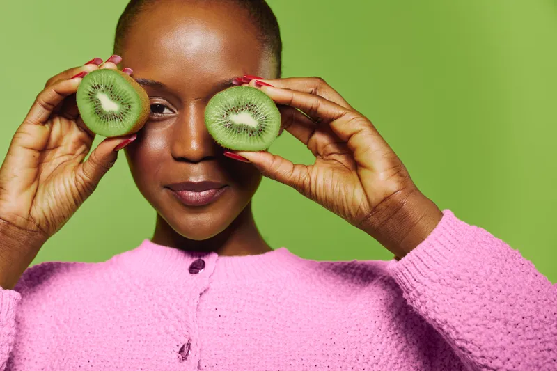 Portrait of a smiling black woman with dark skin holding two halves of a kiwi fruit near her eyes against a green background. Concept of healthy lifestyle and natural beauty.