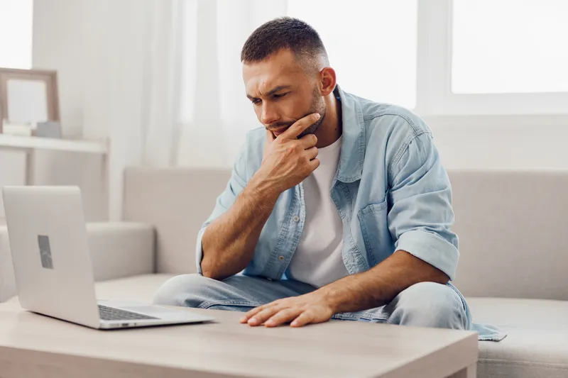 Thoughtful man contemplating decisions while working on a laptop at home. Inspiring and casual workspace complements modern lifestyle and productivity.