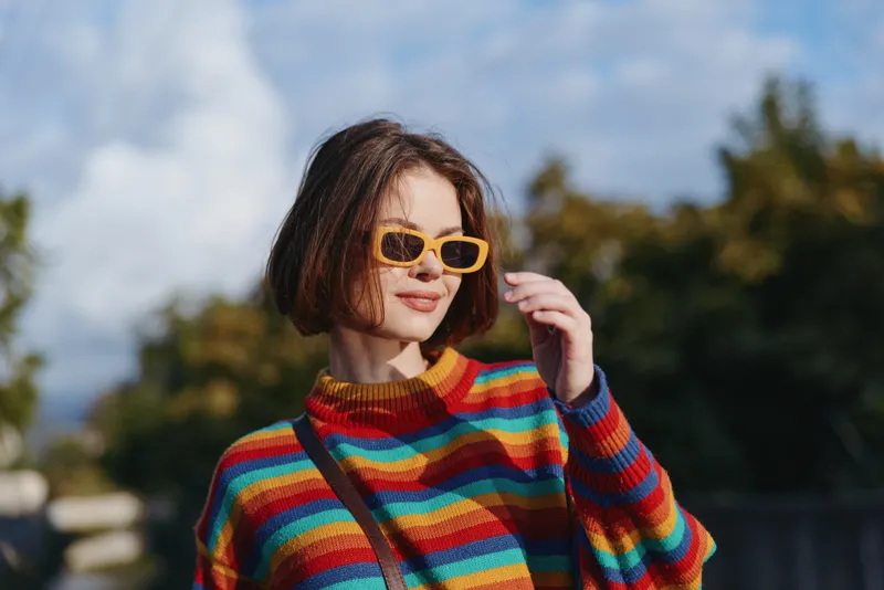 Young woman in a striped sweater and yellow sunglasses, short bob hairstyle, smiling outdoors in sunny park, casual outfit with crossbody bag, lifestyle travel and fashion portrait.