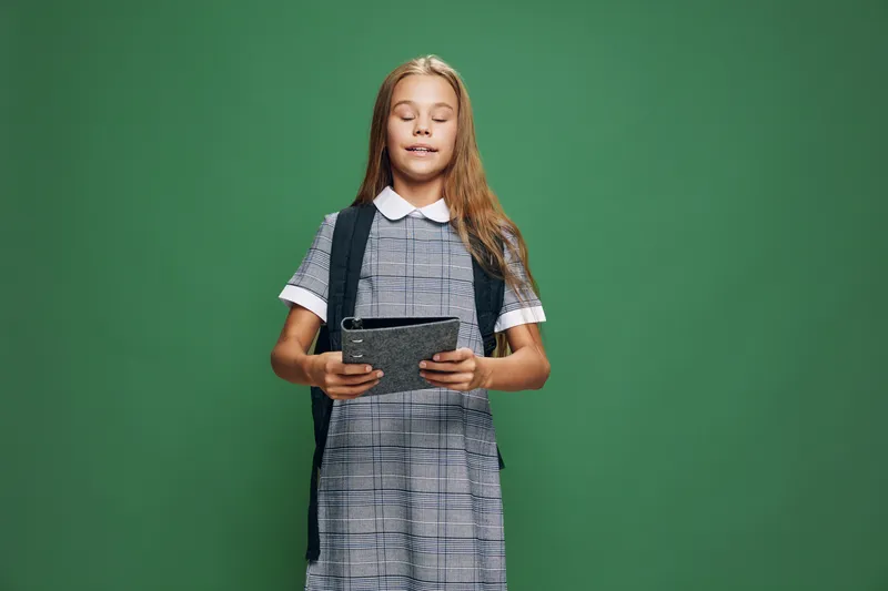 Girl student in checkered dress holding tablet with closed eyes standing against green background for education technology and learning concept.