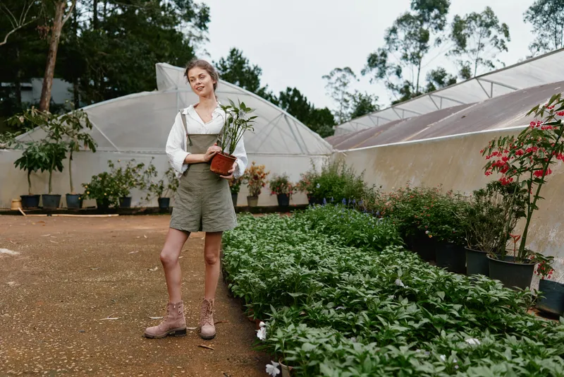 Young woman in a greenhouse, holding a potted flower, dressed in casual summer attire, showcasing a love for gardening and nature