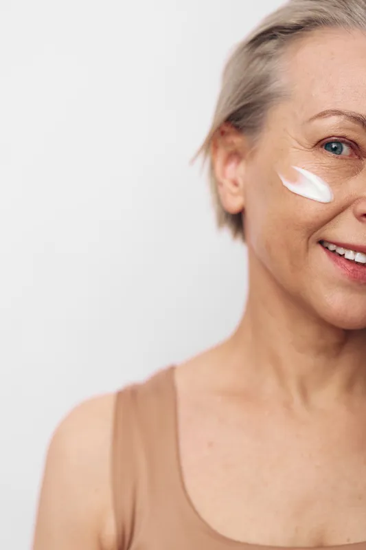 Smiling mature woman with natural skin applies white face cream on cheek, close-up on light background. Concept of skincare, aging beauty, self-care, and healthy lifestyle.
