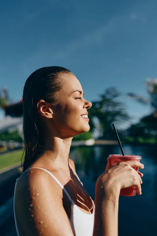 Vibrant outdoor scene shows a smiling woman by a pool enjoying a pink refreshing drink, poolside sunshine, carefree summer mood, lifestyle portrait, candid laughter