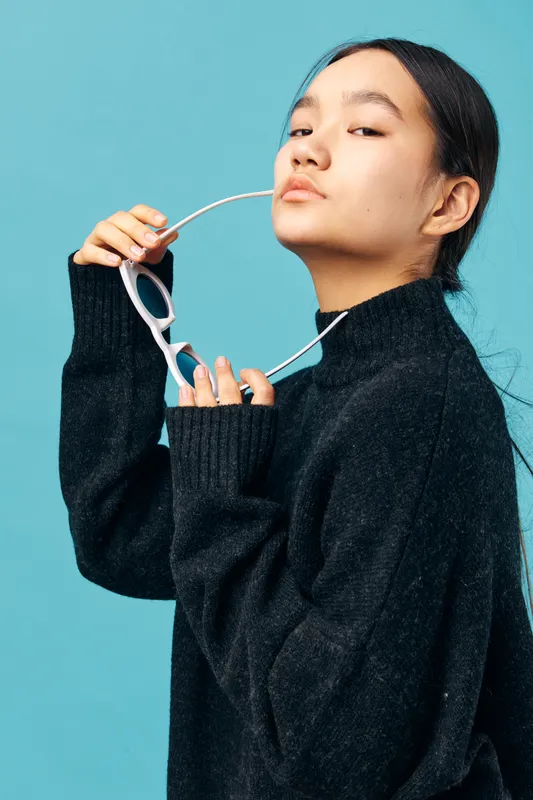 Young woman in black sweater holding white sunglasses with confident expression and pulled back hair posing against blue background in studio for fashion portrait.