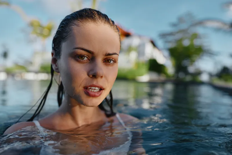 Closeup of a woman in a pool, water on shoulders, sunlit outdoors, wet hair, a candid moment of relaxation and wellness, capturing a refreshing lifestyle scene with natural light and tranquil